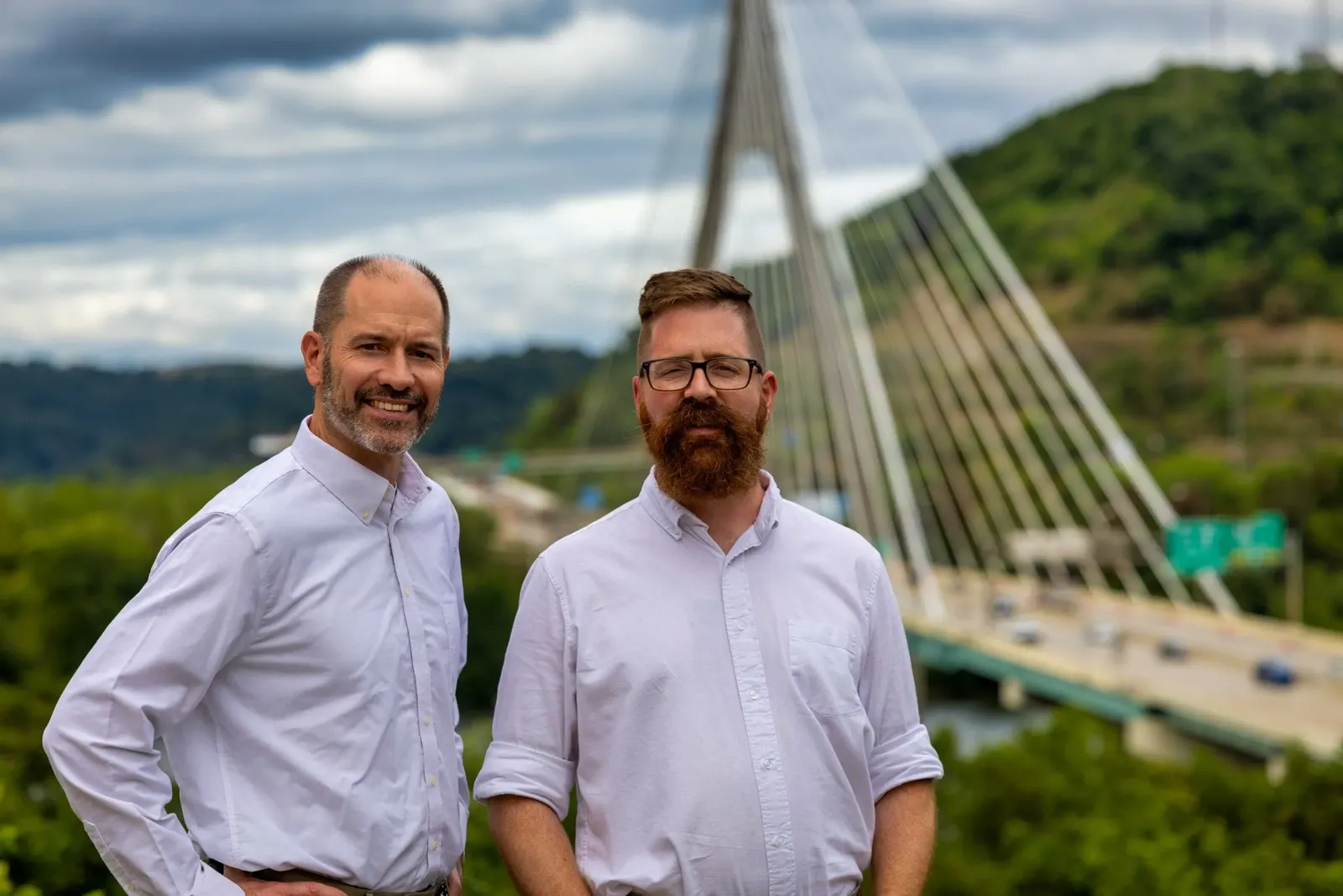 Brian Hough and Martin Watjen standing outdoors above a river valley with a cable-stayed bridge behind them